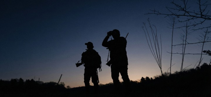 Two VisioGear field testers using handheld thermal monoculars and night vision binoculars for surveillance on a rugged ridge line during deep twilight